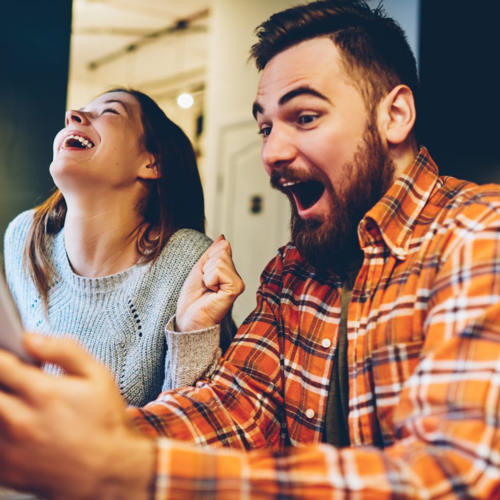A man and a woman shout with excitement as they use the DealDash App on their phones.