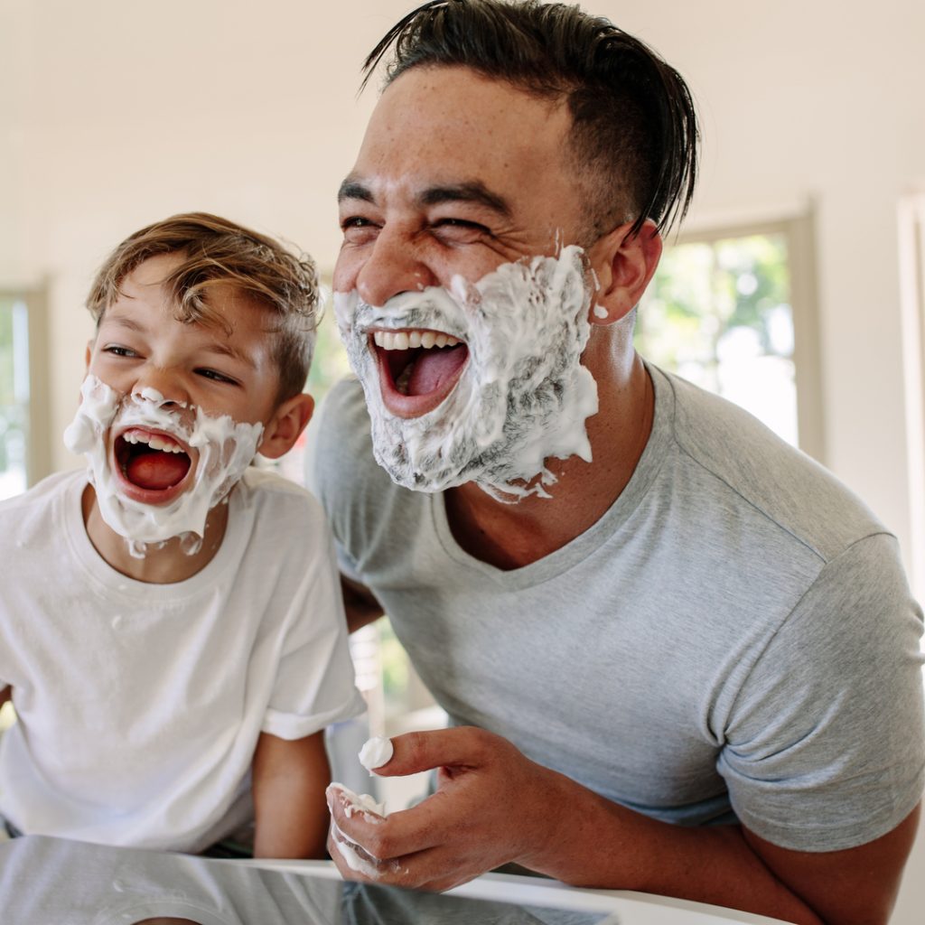 A father laughs while shaving his beard as his son plays with shaving cream.