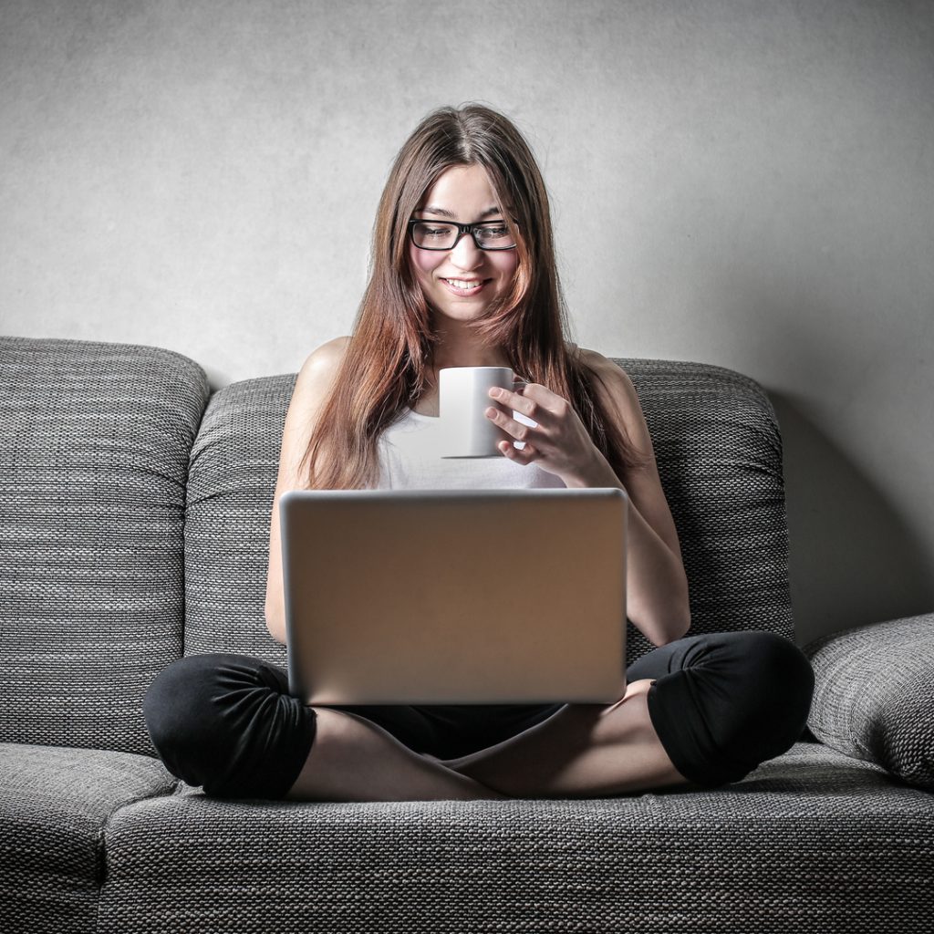 A woman enjoys a cup of coffee while relaxing on her couch and using her laptop.