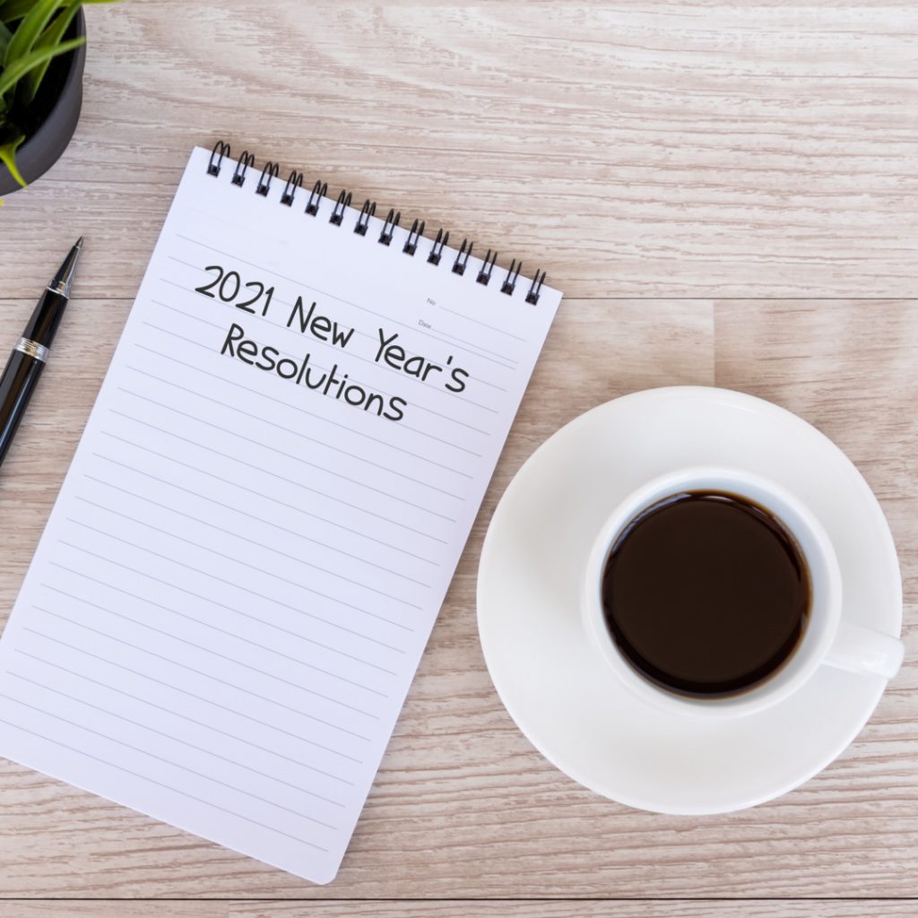 A note pad and a pen wait on a table next to a cup of coffee.  The pad is to be filled out with New Year's resolutions for 2021.
