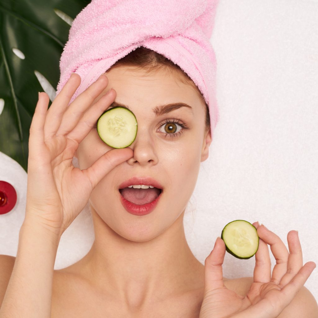 A woman enjoys a facial treatment at a spa and removes a cucumber from one of her eyes to take a peek at the camera.