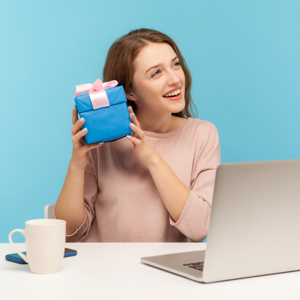 A woman holds up a small wrapped gift while sitting in front of her computer.