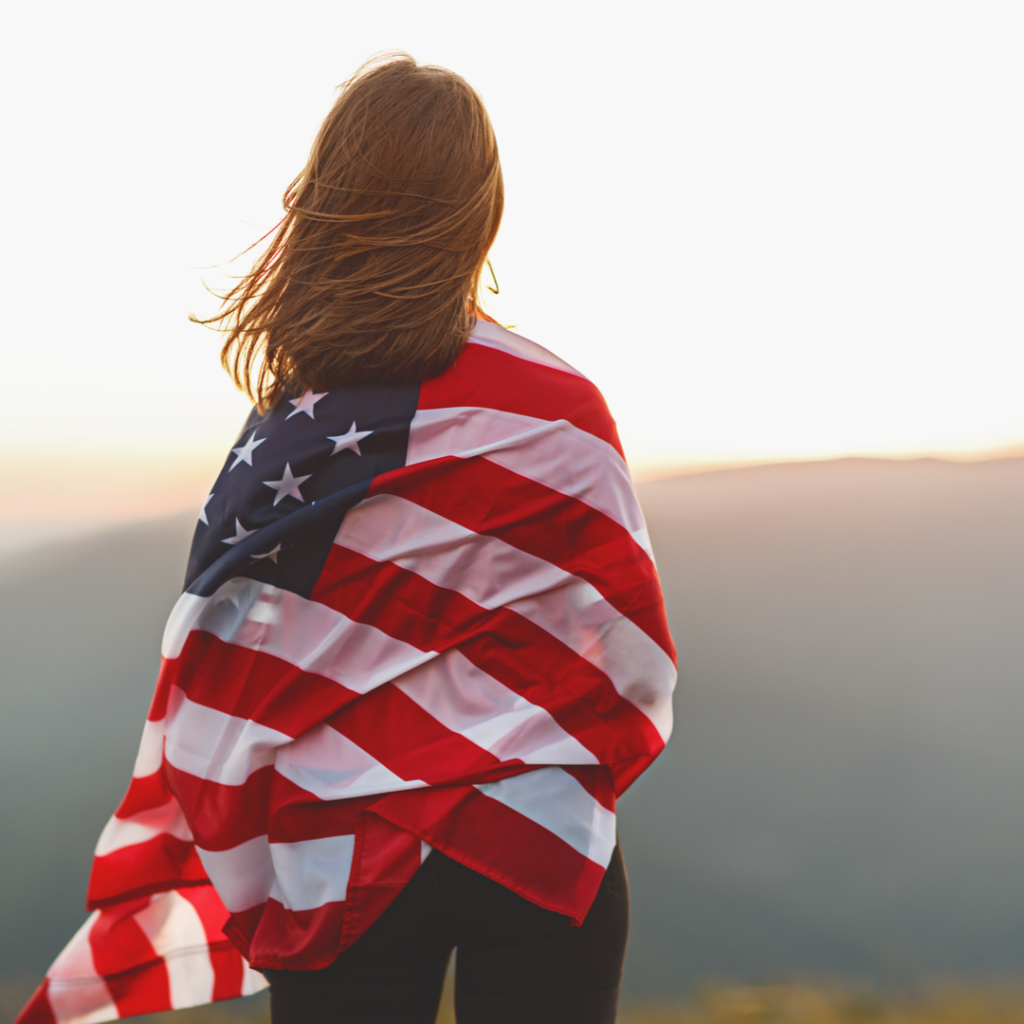 A young woman is wrapped in the American flag and gazes over the countryside.