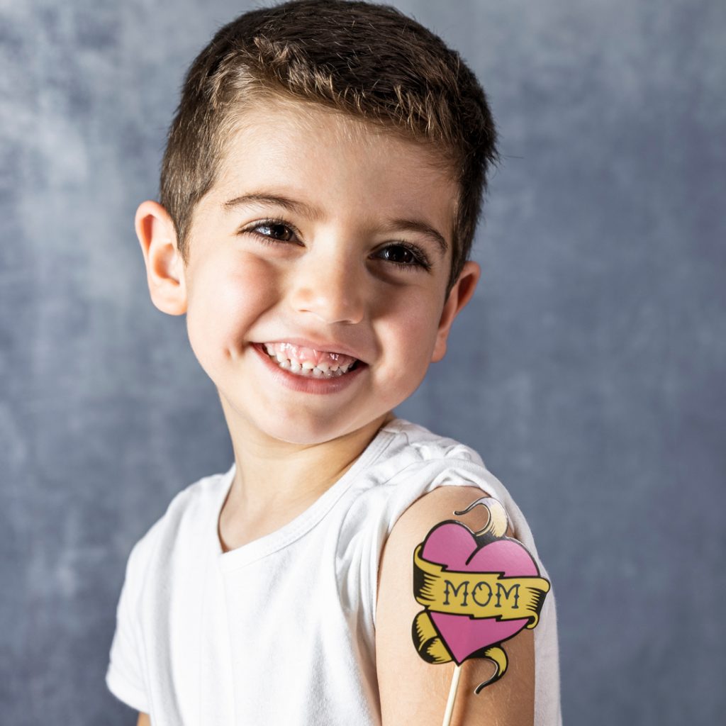 A young boy smiles for the camera with a fake tattoo on his arm that says 'Mom'.
