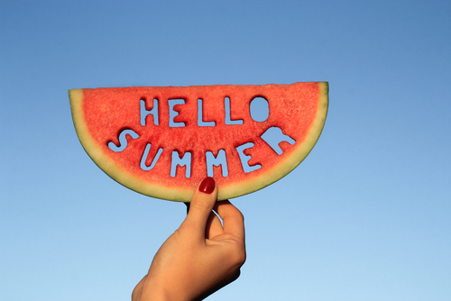 A watermelon with the words 'hello summer' cut out of it is lifted in the air.